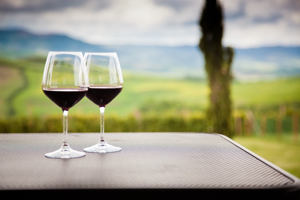 two glasses of red wine on a table with green Tuscan hills in the background